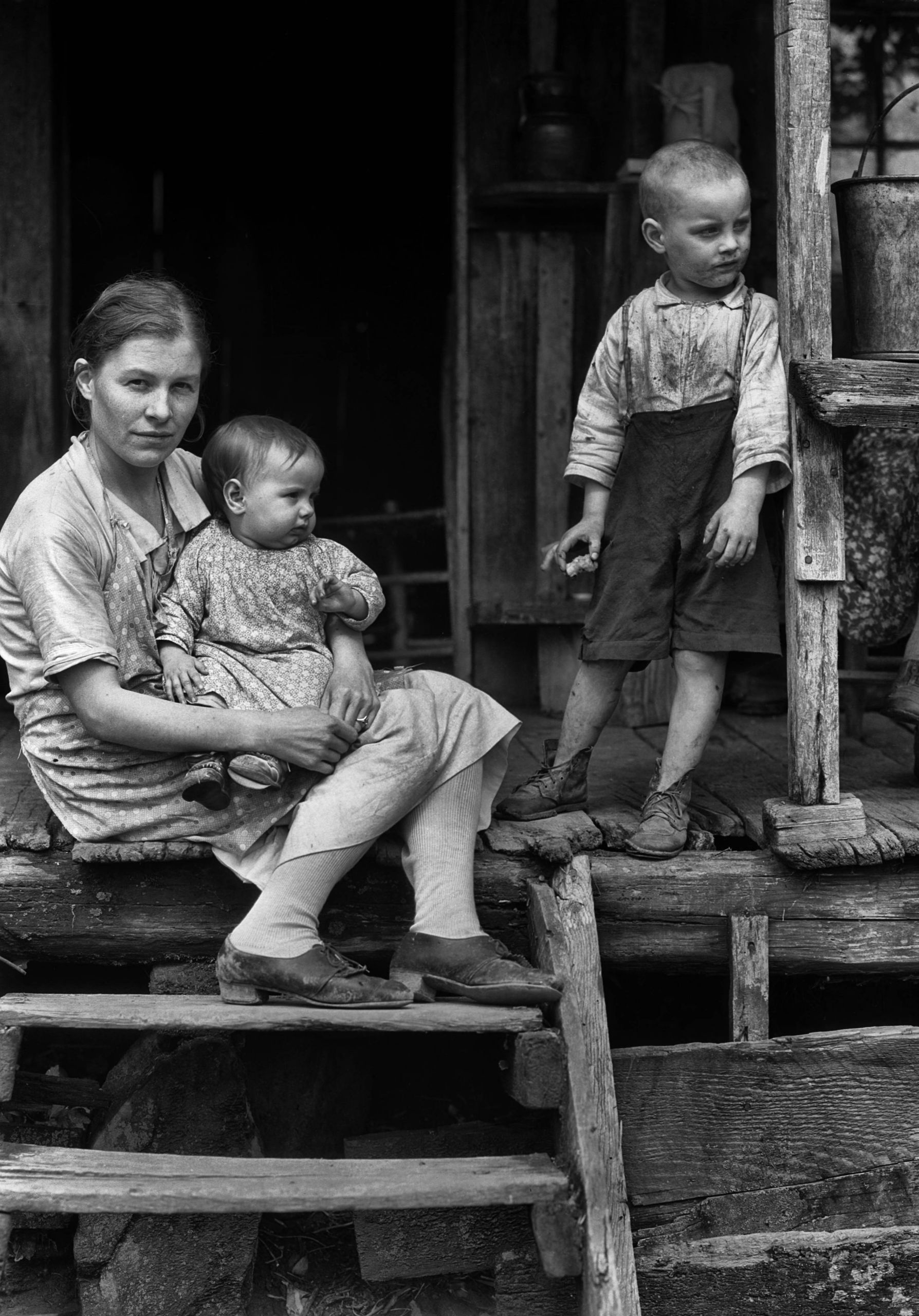A mother with a baby seated on a porch while a young child stands nearby.