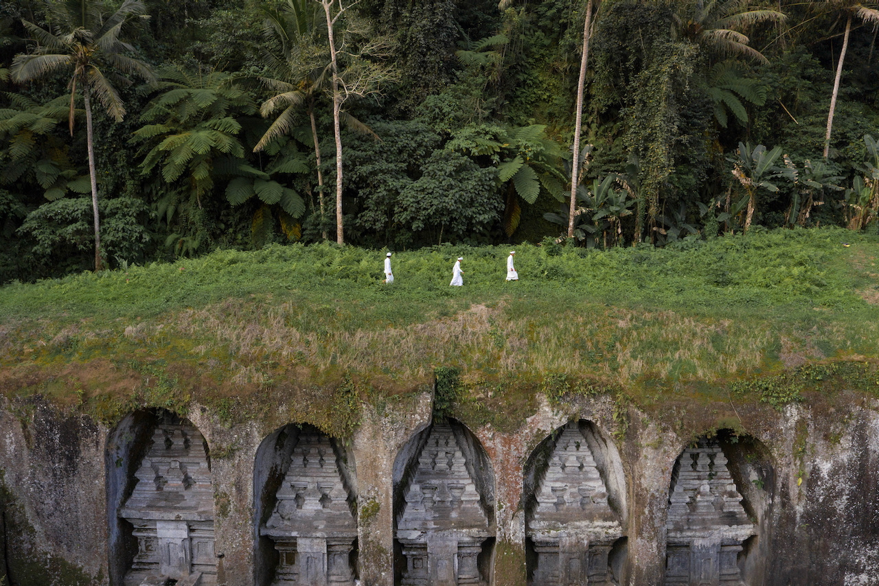 Three small white figures walk above a row of carved stone niches in the jungle, Bali