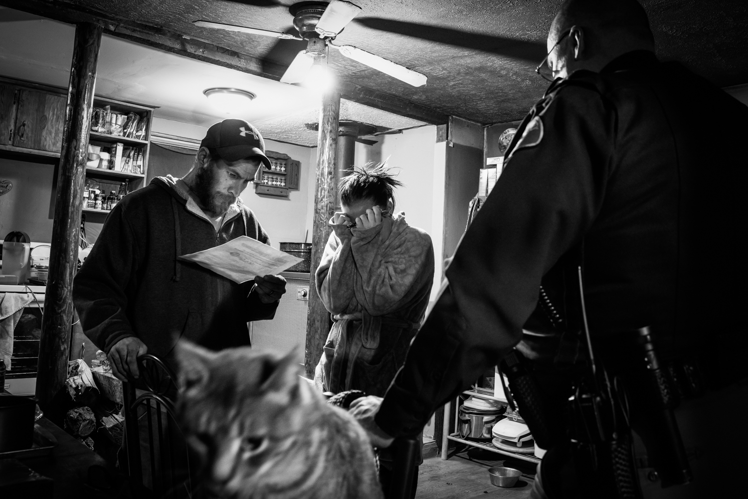 A man reads a search warrant while a woman covers her face, a police officer stands in the background, Nebraska